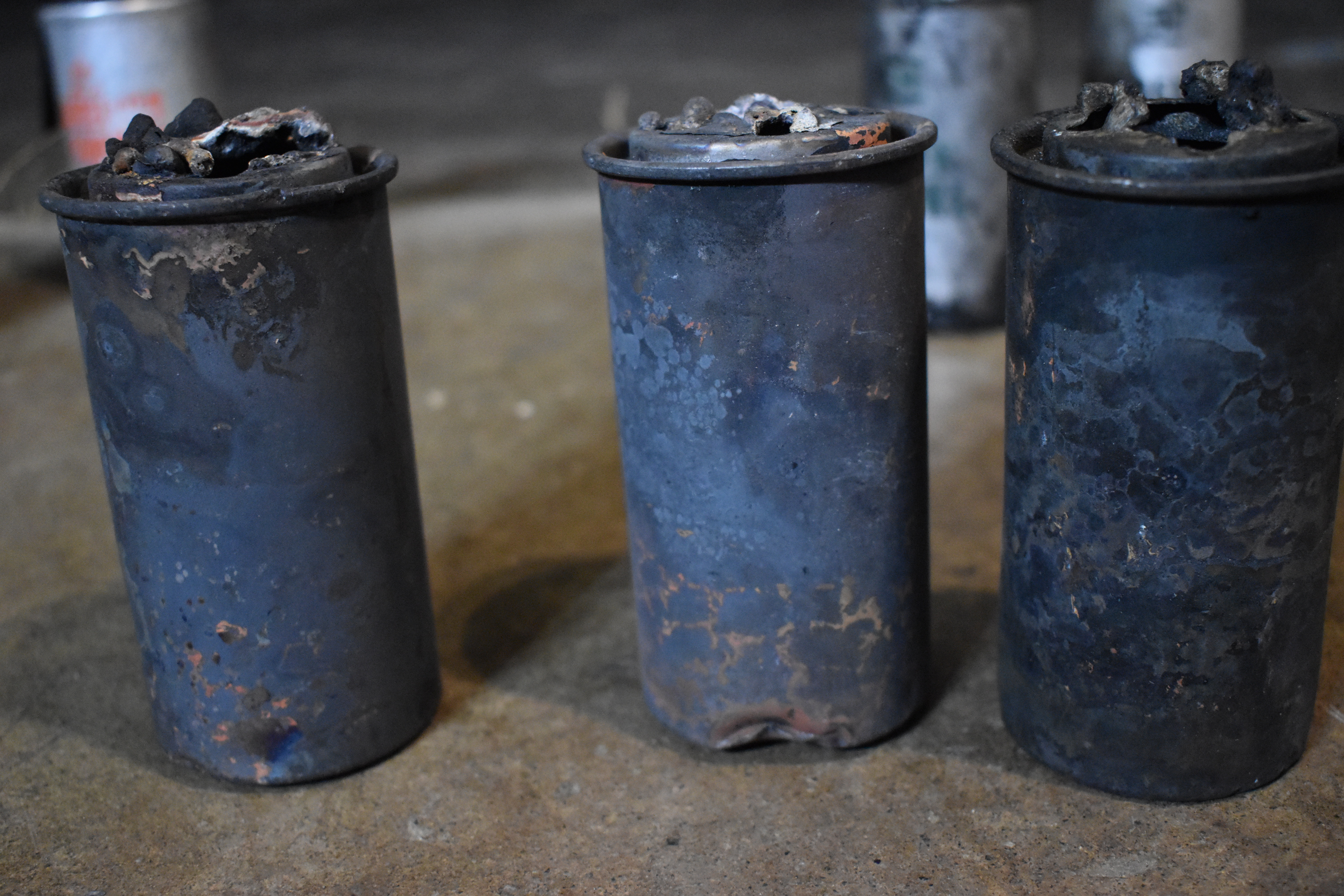 Three canisters of HC stand in the foreground of the image, the background yields three other canisters of various chemical agents. The HC canisters are cylindrical with riveted tops resembling a paint can lid. Their markings have burned away with the heat of their ignition. Metal along the top has melted, peeled and been blown outwards. Their rough rounded edges border holes into the center of the canister. Chemical reactions have changed the color of the metal to dark grey, rust brown, and a navy blue. The canisters are dented from impact and appear to have lost any smoothness they may have once possessed.