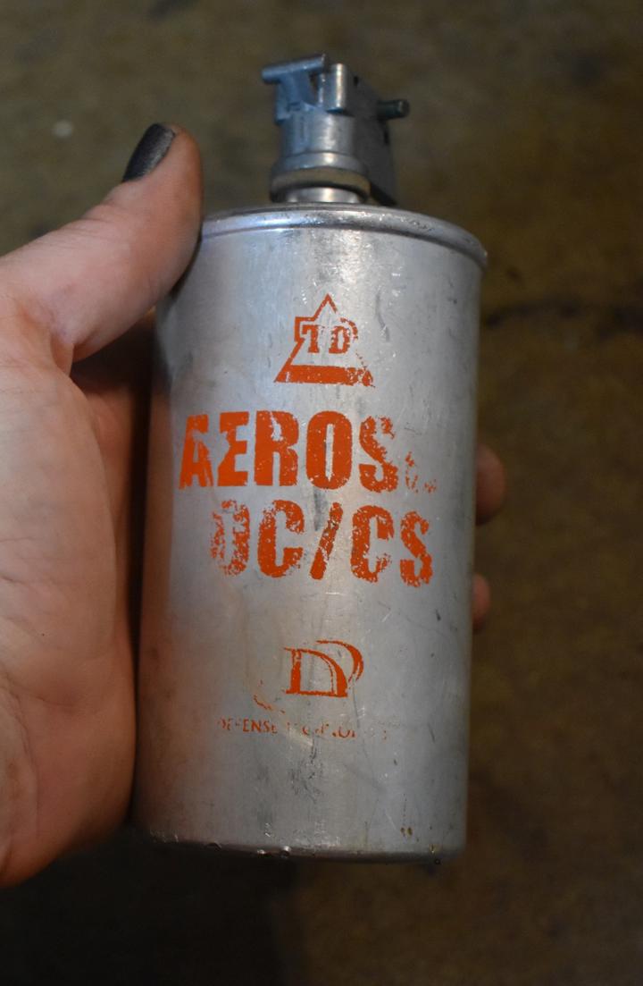 A hand holding an old silver can with orange writing that says `TD Aerosol OC/CS Defense Technology.` the canister is an aluminum cylinder, with a lip at the top of the tubing, and a spring and fuse mechanism that makes the canister resemble a grenade.