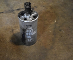 An old silver spent grenade can is pictured with white writing that says `CM Riot control, CS, Defense Technology` the canister has four relief holes machined into the top of the canister, and a spring mechanism that appears to be the trigger mechanism sits in the exact center of the lid.