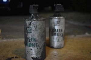 Two old aluminum smoke grenade canisters are pictured with green writing that says `CM Green Smoke Defense Technology`. Both still retain their black plastic trigger mechanism covers, but neither have their fuse rings. Both have been used. The 6 inch tall, nearly two and a half inch wide aluminum canister has a crimped smooth lid at the top, securing the fuse plug in place.