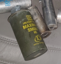 An old green smoke grenade canister with yellow writing that says `CM military style maximum smoke HC Defense Technology` sitting on a car seat with other munitions behind it. The canister is nearly 2.5 inches in diameter and 5.5 inches long. The black plastic trigger cap and lever have come off during use. It's covered in a thin layer of dust or residue.