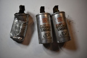 Three old aluminum canisters with orange writing that says `CM Military Style Orange Smoke Defense Technology` are pictured. The each retain their black plastic trigger mechanisms but no fuse rings. They are over 5 inch long aluminum metal cylanders with a lipped lid securing the fuse plug, which has been smoothly crimped the the cylinder's body. The canisters pictured are damaged, dented, and it appears one has been run over.