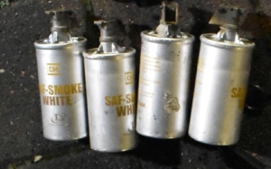 four old aluminum grenade canisters are pictured with yellow writing that says `CM SAF Smoke White Defense Technology` they each retain parts of their black plastic trigger mechanisms but non of them are pictured with their levers or their fuse rings. They're cylanders that resemble a pringles can with smoothly crimped lids to secure the trigger mechanisms in place. Metal support joysts secure the trigger mechanism further, to prevent shifting or bending.