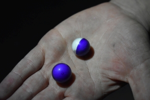 handle holding two pepper balls in an open palm, one is purple, one is half purple half white. The Pepper Balls are made of a smooth plastic. Each half is capped together like a plastic easter egg. Inside, compacted white powder is just barely visible beneath the purple transparent caps.