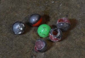 five half-black half-red pepper balls, three broken open two intact, surrounding a green marking pepper ball are pictured on the ground. They're plastic balls, round and smooth, made up of two parts that snap together like an Easter Egg. Inside the intact ones we can just barely see compacted powder. Outside the broken ones, they are covered in a white dust.