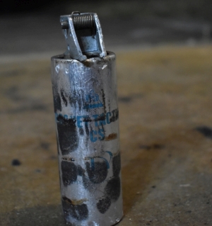 old small aluminum grenade canister with blue writing that says `TD Pocket Tactical CS`, the canister is a roughly 5 inch long cylinder of about an inch and a half in diameter. The black plastic fuse trigger has broken off to reveal the metal fuse trigger components and cannister plug. The canister is covered in scorch marks and scuffs from its use in the field.