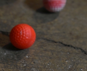 Small dimpled spheres that resemble golf balls are pictured on the ground. Their paint colors vary, but the ones pictured are red and a gradient of white to yellow to Orange to pink and red. These dimples function to make the spheres noticeably more aerodynamic when propelled. They're made of a smooth plastic like substance that appears hard to the touch. Much like a smaller golf ball.