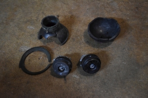 components of a black plastic, ball hand grenade, about softball sized, shaped like a ball, but broken and split apart. The trigger mechanisms popped off, missing their black plastic trigger caps. A gasget is also pictured that seems to fit between the two halves of the grenade.