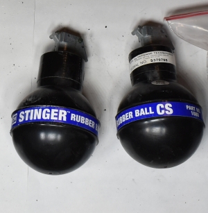 two un exploded stinger rubber ball CS grenades black with blue label with white writing on it that says Stinger Rubberl ball CS, background is white paper with shadows