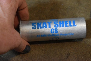 The SKAT Shell is pictured on concrete ground, held in place by a person's hand with gun metal grey nailpolish. The shell is a long cylindrical tube with a lip at the bottom, similar to a shotgun shell. It's made of what looks like stainless steel or aluminum. Silver in color. Faint scratch lines along the metal that are more known than felt. It looks to be about 5 inches long. Stamped on the shell in a thin paint designed for metal cans like soda pop and beer are the words; `Defence Technology, SKAT shell, CS, 40 millimeter multiple projectile.` SAFARILAND is written at the bottom with a part number and expiration date.