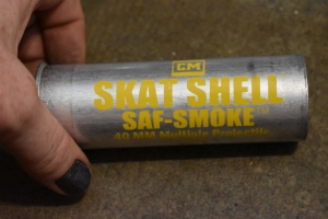 An aluminum cylinder of roughly 5 inches long and 40 millimeters wide is pictured on a smooth concrete floor. The cylinder is stamped with metal paint that reads; `CM, Skat Shell, Saf-Smoke, 40 millimeter multiple projectile` The shell has a Defence Technologies logo on the bottom of it, and a warning message below it indicating this product is for authorized personnel only. Beneath this it says `Safariland` above an experation date and a product number. The shell itself resembles a large shotgun shell, lipped at the bottom of the cylander.