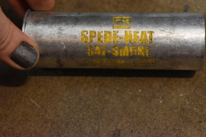 A long cylinder is pictured on the ground with smooth concrete beneath it. The cylinder is roughly 5 inches long and made of a shiny silver metal, such as aluminum or steel. There is a lip at the bottom like one would find on a shotgun shell. Stamped on the metal are the words `Defence Technology, Spede-Heat, Saf Smoke, 40 millimeter long range - 150 yard, 40 millimeter single projectile.` The lettering on the shell has been damaged and is largely illegible.