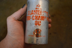 A hand is pictured holding a old aluminum, silver canister, with orange writing that says 'TD flameless tri-chamber oc Defense Technology' A mechanism at the top made of black plastic appears to be some kind of grenade trigger. Like an old bug bomb cannister. It's lipped at the top of the aluminum cannister, where the lid is crimped over the canister, shaped like a cylinder. The metal is worn and scraped and dented.