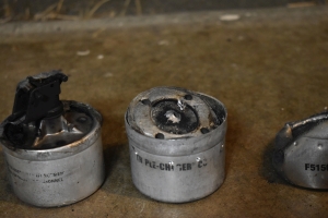 three parts of a Defense Technology Triple Chaser CS Grenade set next to each other with black scuffed text identifying the canisters. The canisters are aluminum, and designed to stack together, so there's a flared aluminum seat above 2 of the 3 canisters, though this is not shown well here as one of the canisters seems to have fallen apart and the other appears to have nearly completely melted, the flared aluminum seats usually resemble a small cookie tin without a lid. On the third piece, a lid is crimped on smoothly with a trigger mechanism within the lid. The trigger mechanism here is shown with the plastic trigger still covering the metal trigger mechanism. Around the trigger mechanism, 4 holes are machined into the trigger lid surrounding the trigger machanism itself. Inside the seats of the two canisters without triggers, 4 machined holes set into their lids around the center of the canister, and a 5th hole in their centers. Though this is not shown for the melted canister.