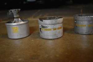 three parts of a Defense Technology Triple Chaser SAF Smoke Grenade set next to each other with yellow text identifying the canisters. The canisters are aluminum, and designed to stack together, so there's a flared aluminum seat above 2 of the 3 canisters, resembling a small cookie tin without a lid. On the third piece, a lid is crimped on smoothly with a trigger mechanism within the lid. The trigger mechanism here is shown as metal components missing pieces due to being used. Around the trigger mechanism, 4 holes are machined into the trigger lid surrounding the trigger machanism itself. Inside the seats of the two canisters without triggers, 4 machined holes set into their lids around the center of the canister, and a 5th hole in their centers.
